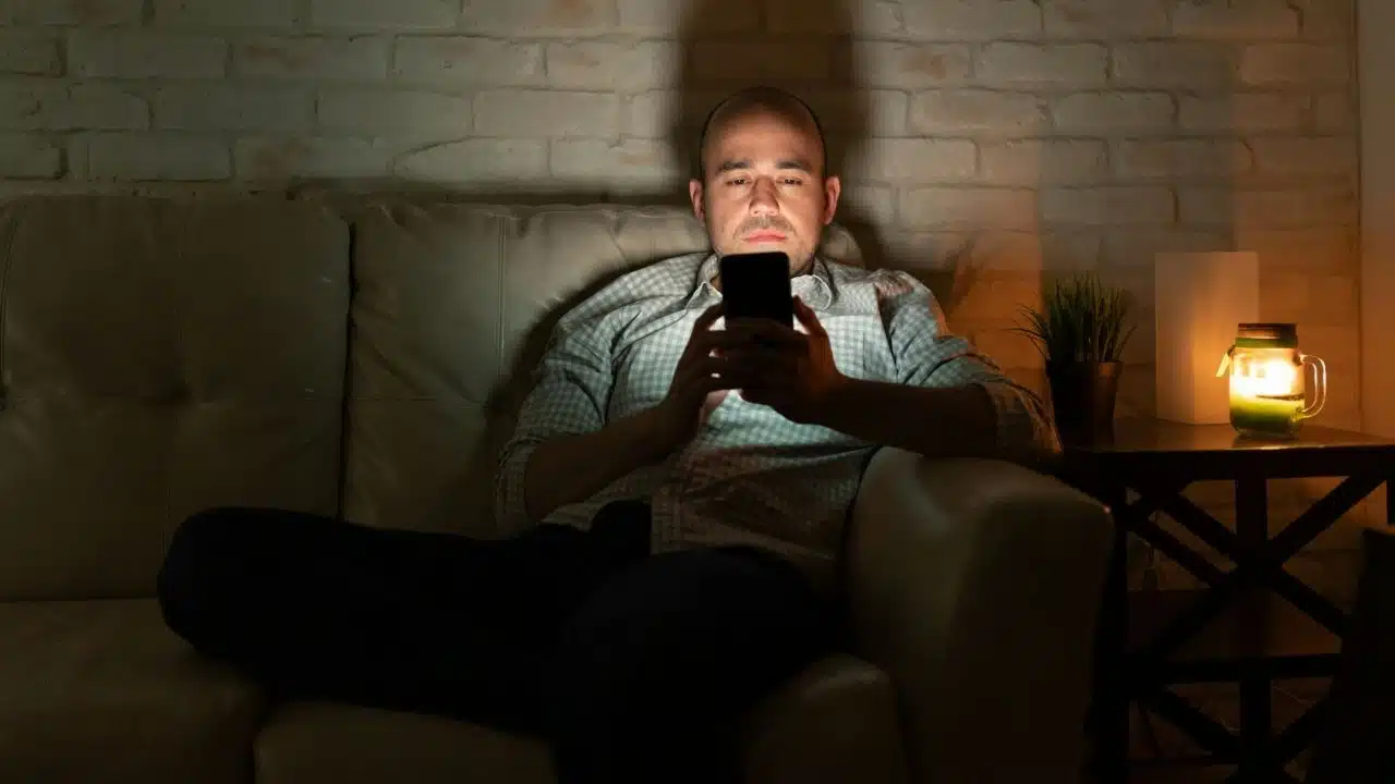 A man sits on a sofa in a dark room looking at his glowing smartphone screen.