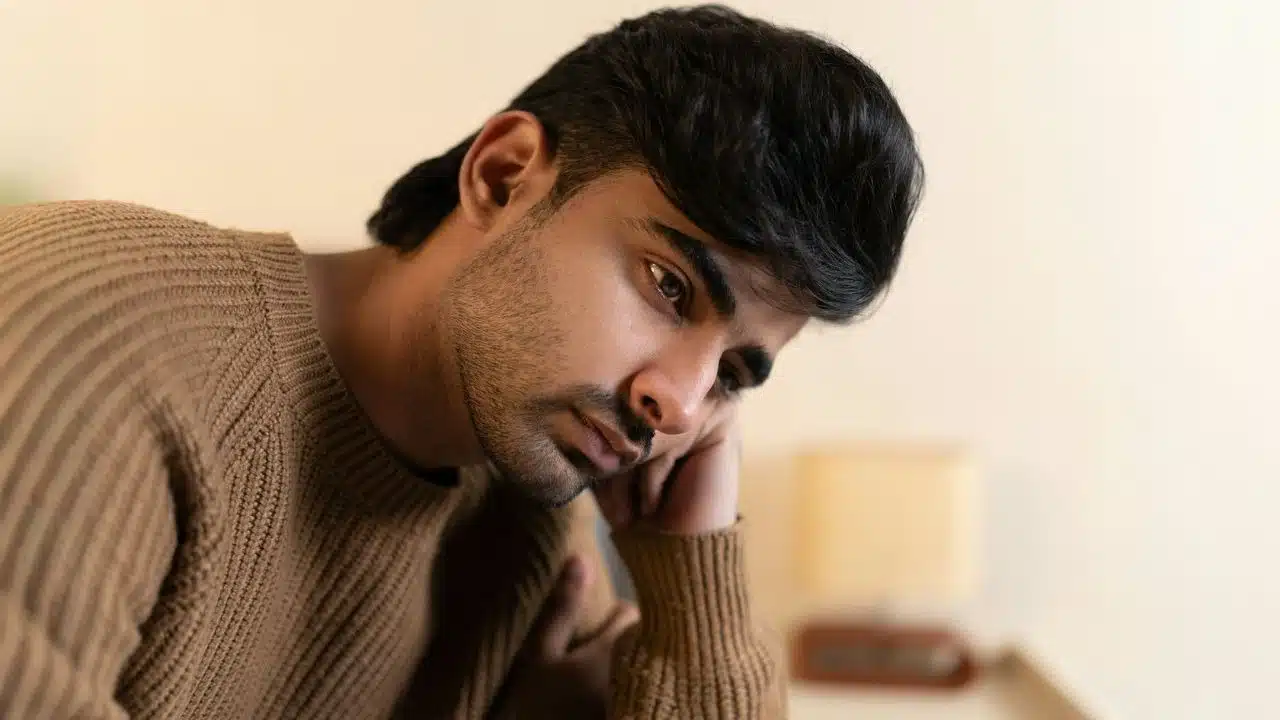 A man in a brown ribbed sweater rests his head on his hand and stares.
