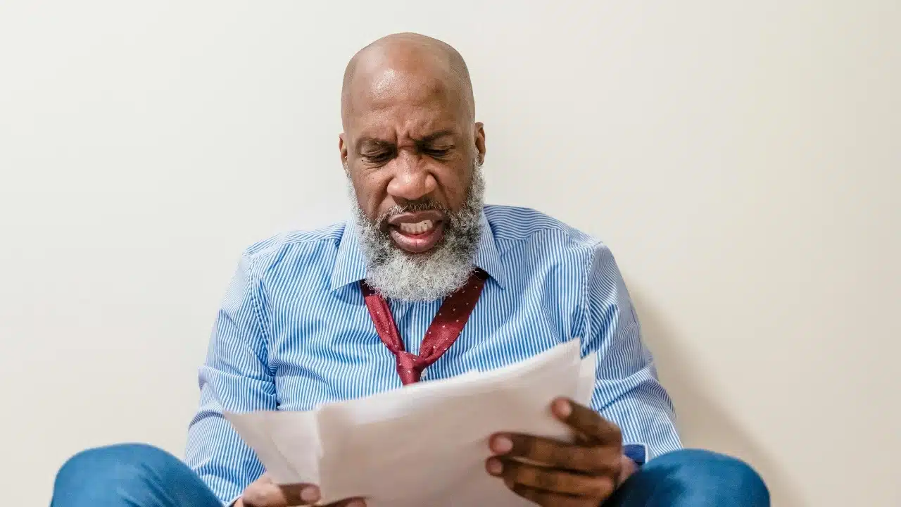 A man with a grey beard sits on the floor looking at papers with frustration.