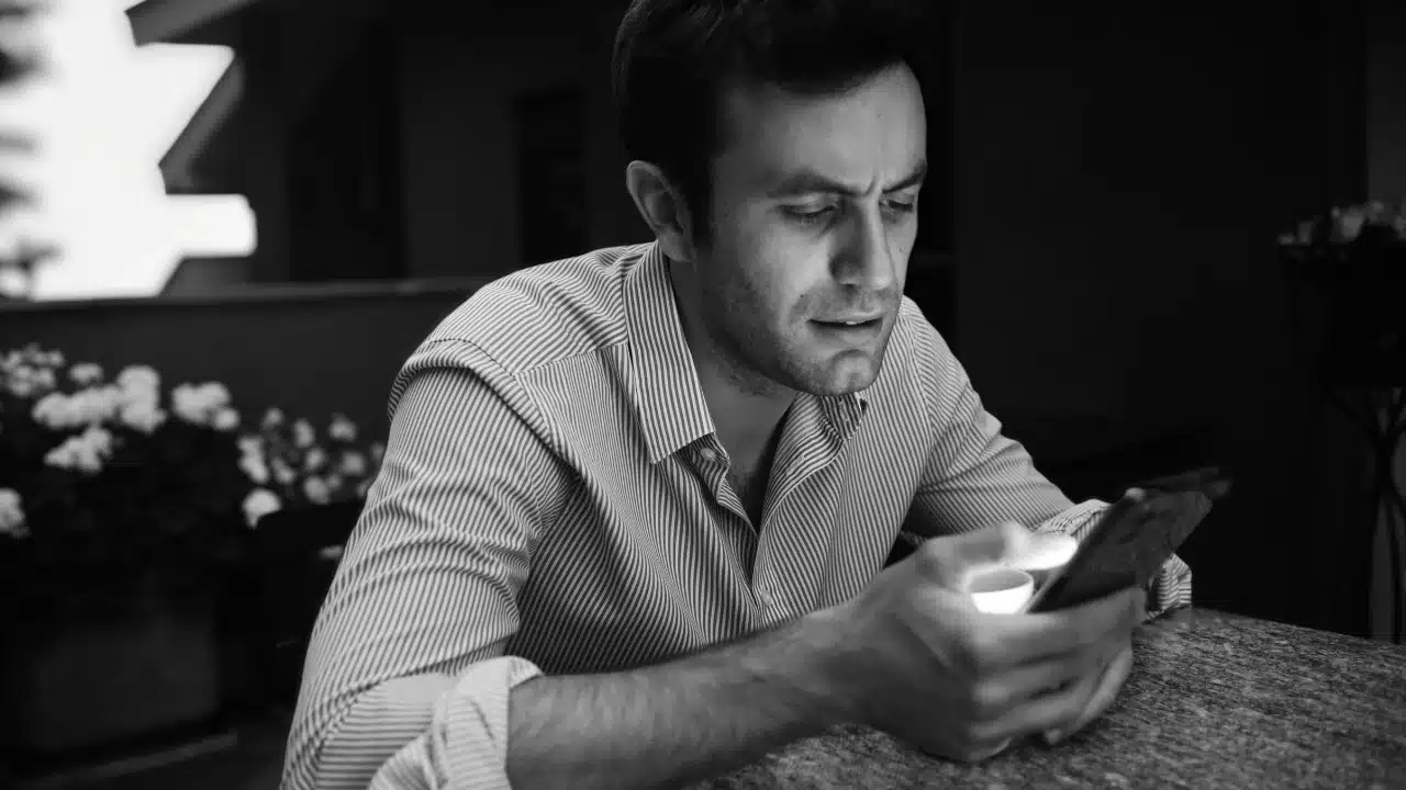 A black and white photo of a man looking down at a glowing smartphone screen.