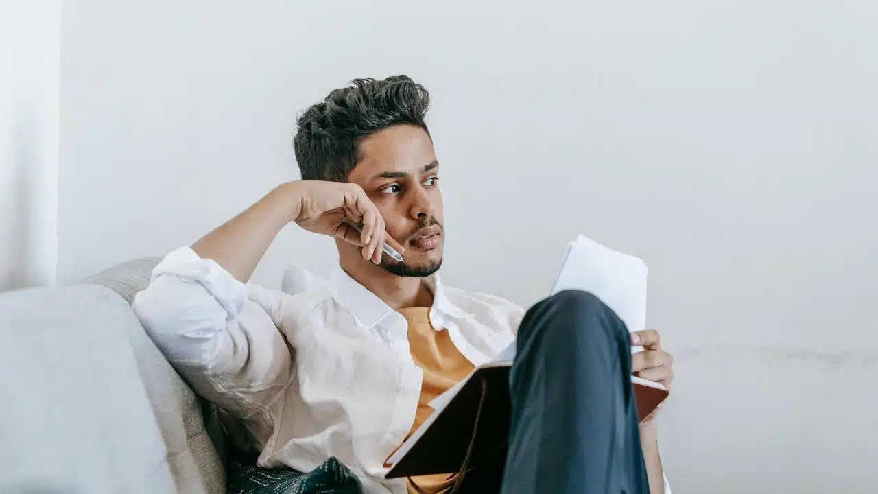 A man sitting on a couch, holding a notebook and looking thoughtfully to the side