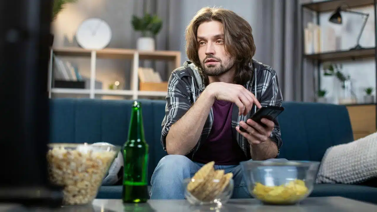 A man sits on a couch with a phone, surrounded by snacks and a green bottle.