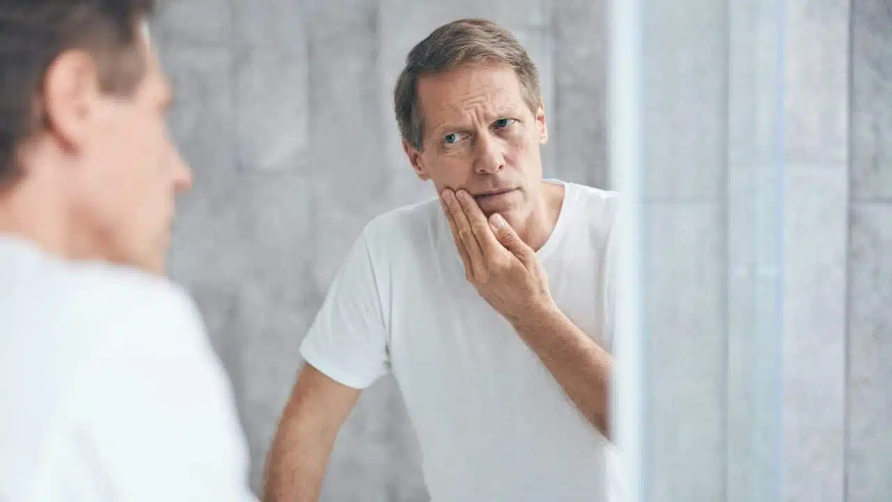 A man in a white t-shirt looks into a mirror while touching his face with concern.