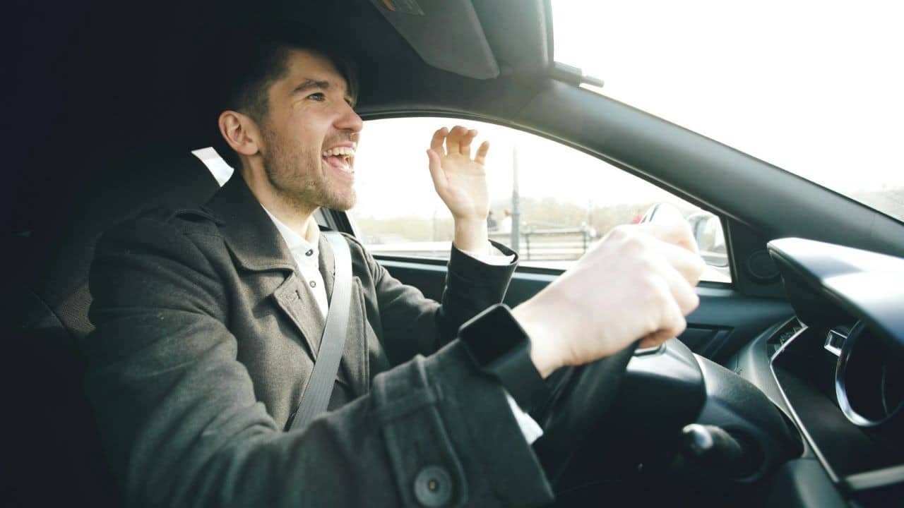 A man in a dark coat shouts and gestures with one hand while driving a car.
