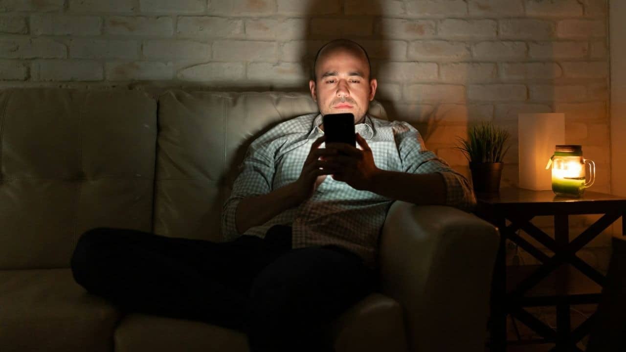 A man sits on a sofa in a dark room looking at his glowing smartphone screen.