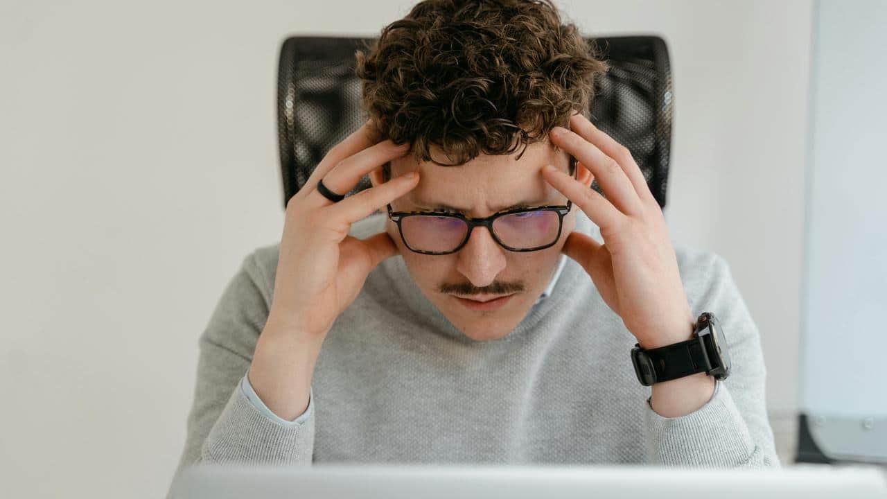A man with glasses holding his head while looking stressed at a computer.