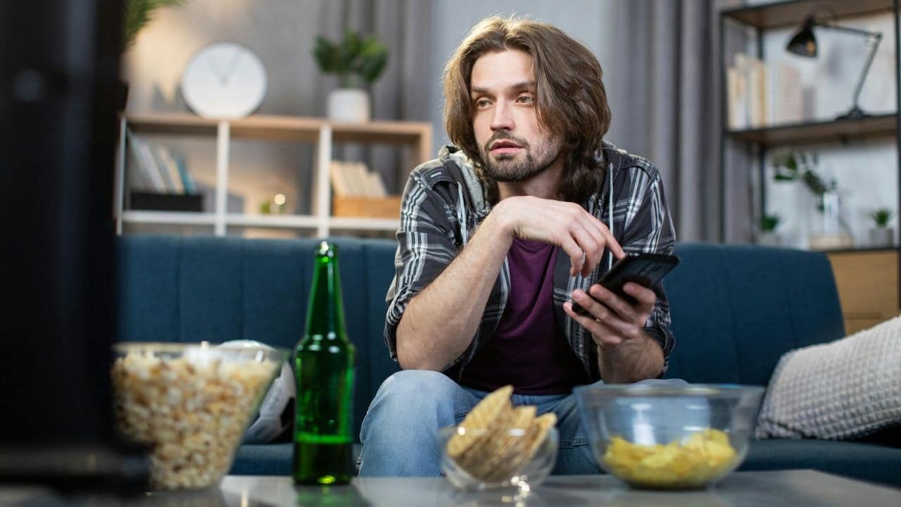 A man sits on a couch with a phone, surrounded by snacks and a green bottle.