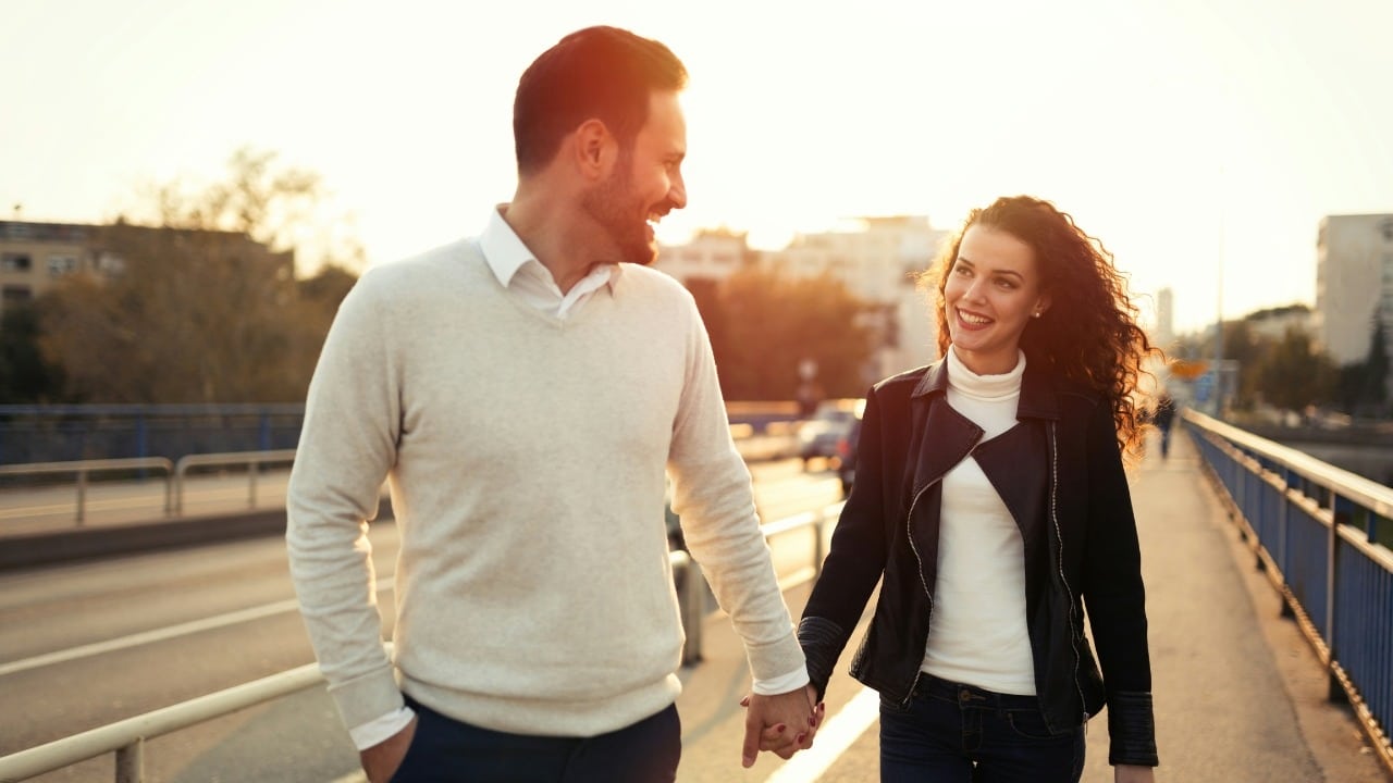 A man and woman walk across a bridge holding hands while smiling at one another.
