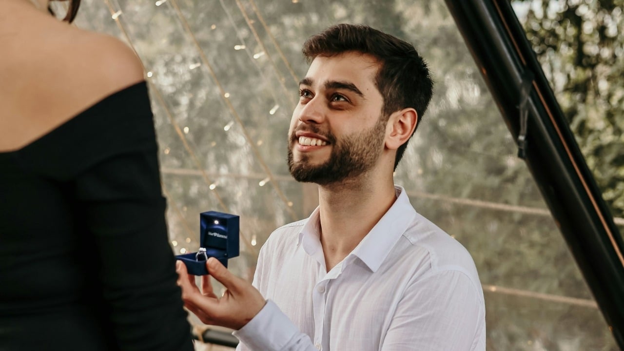 A man kneels and holds an open blue ring box toward a woman in black.