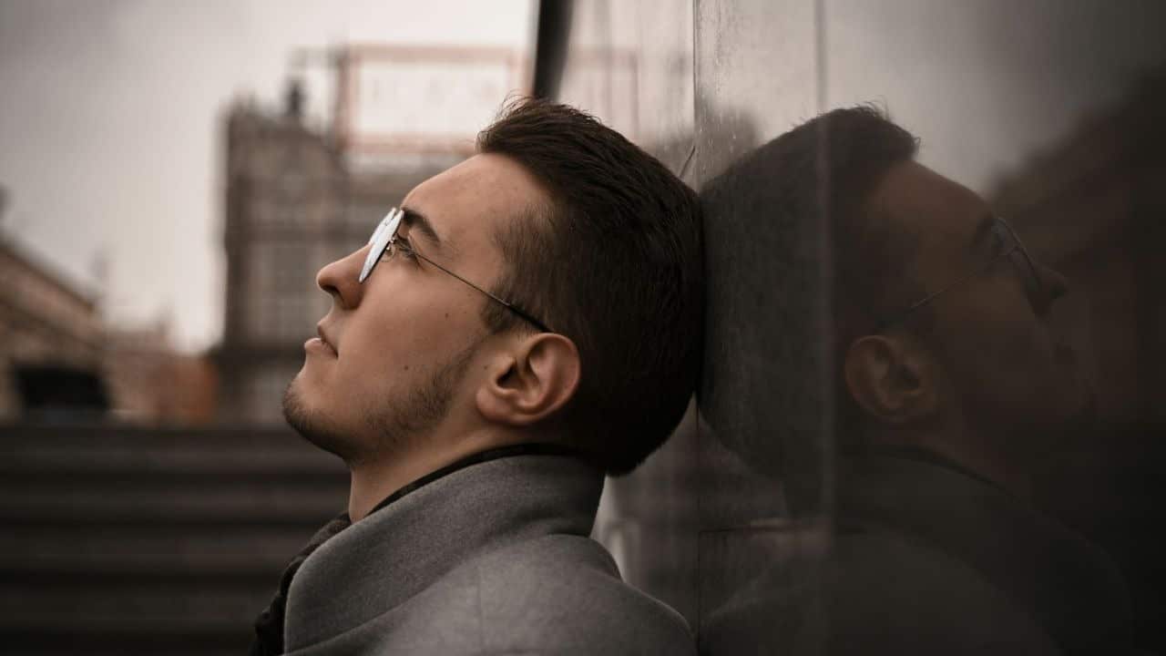 A serious man with glasses and a beard is leaning his head against a stone wall, looking up.