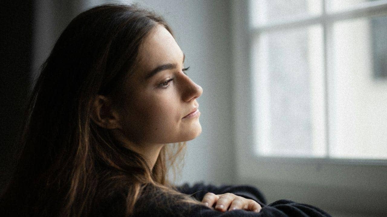 Woman with long brown hair looking out a bright window with a thoughtful expression.