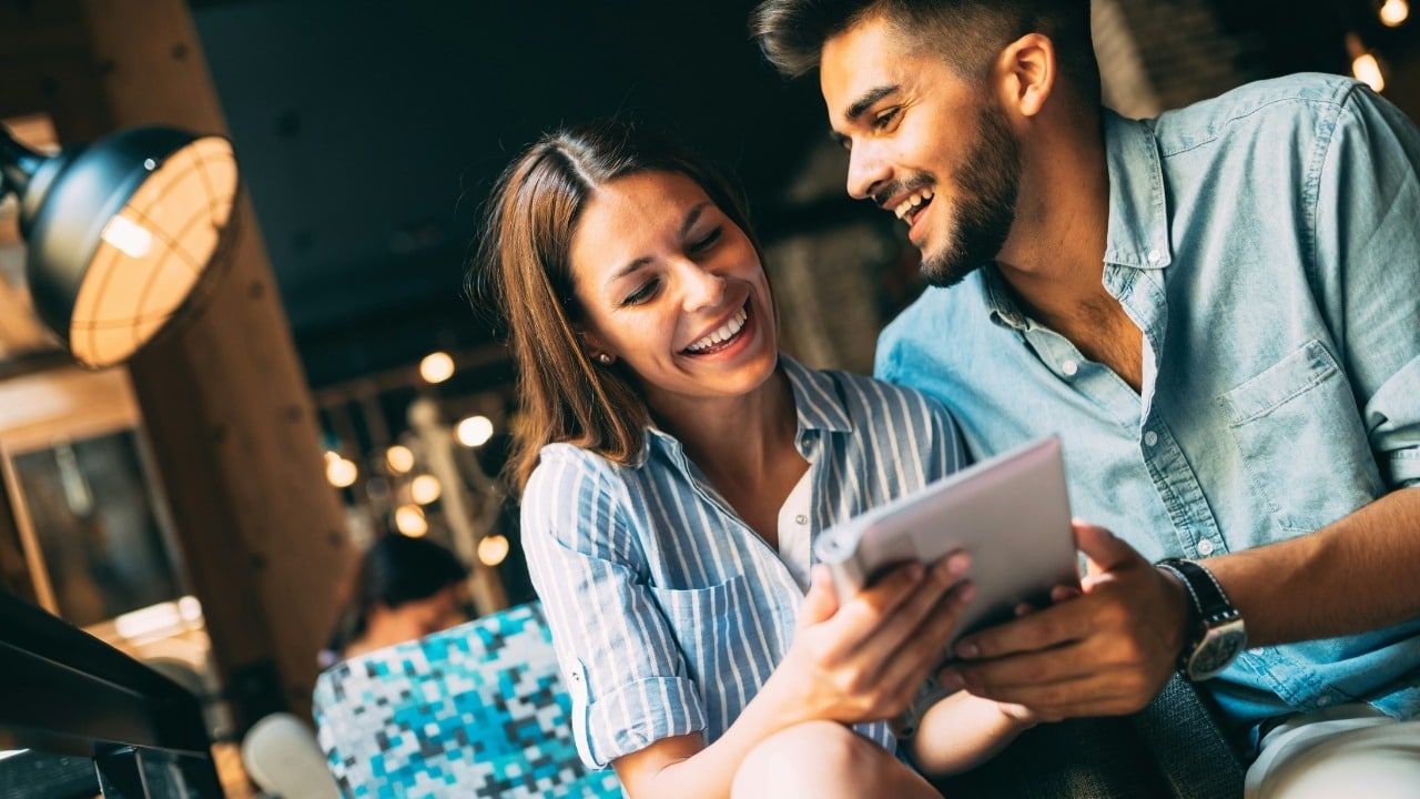 A man and woman laugh while looking at a tablet together in a dimly lit cafe