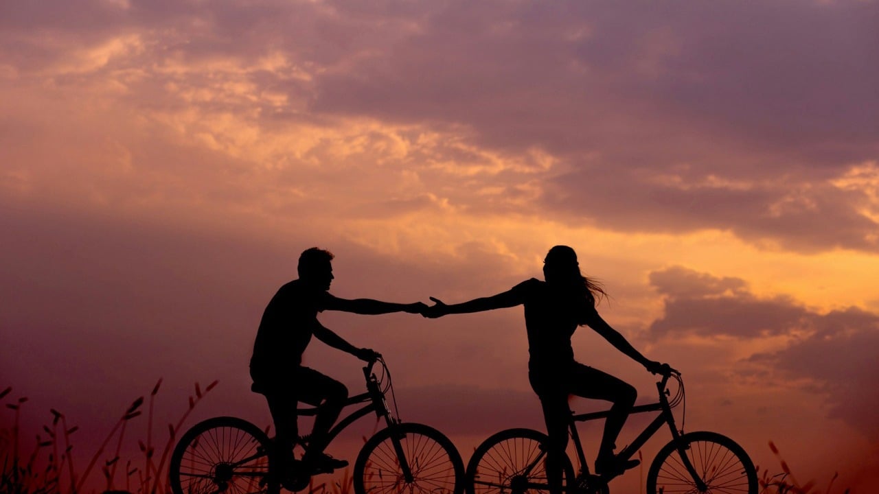 A woman on bike reaching for the man's hand behind her also on the bike.