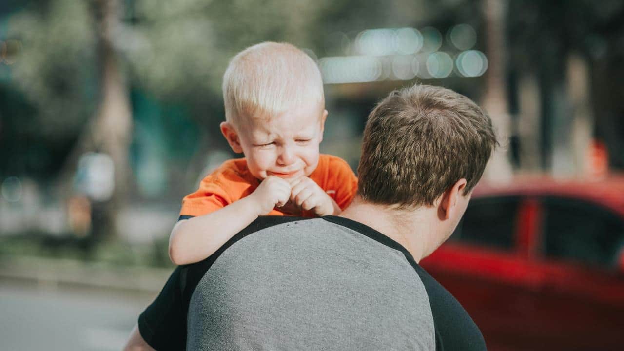 A man carrying a crying child on his shoulder outdoors.