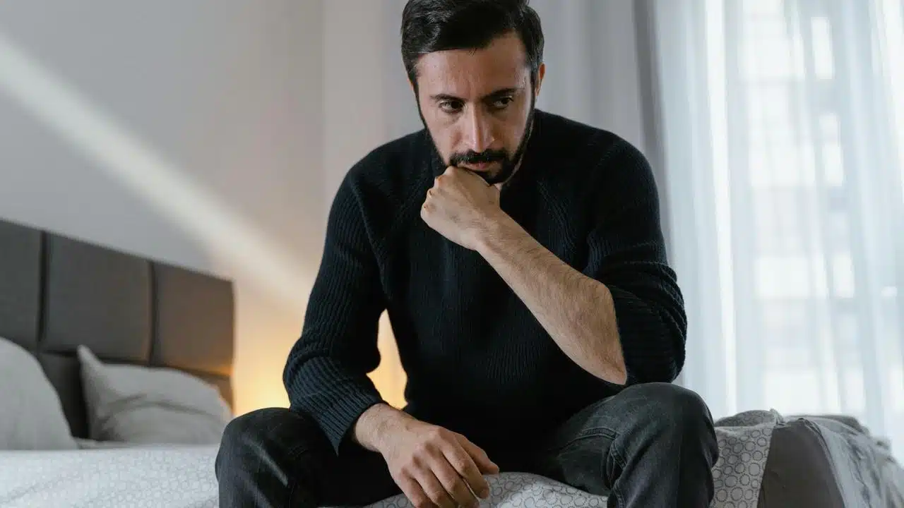 A man in a black sweater sitting on a bed with a pensive expression.