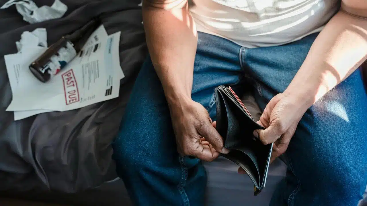 A man holding an empty wallet while past due bills and a bottle sit nearby.