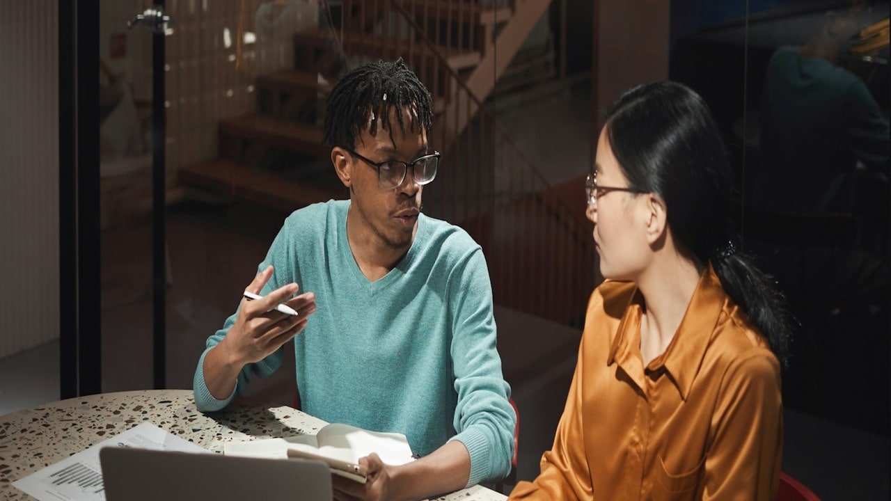 A man holding some papers is talking to a woman sitting next to him.