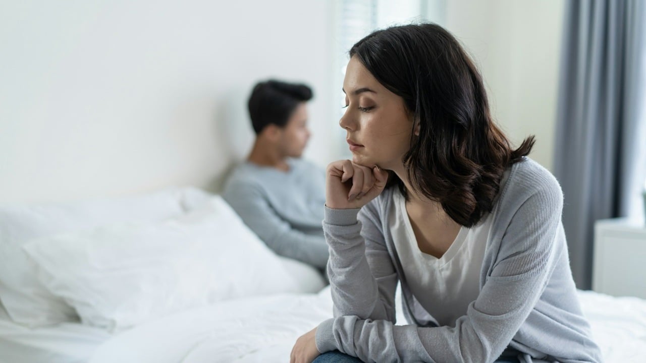 A woman sitting on a bed’s edge looking upset while her partner sits on the bed in the background.