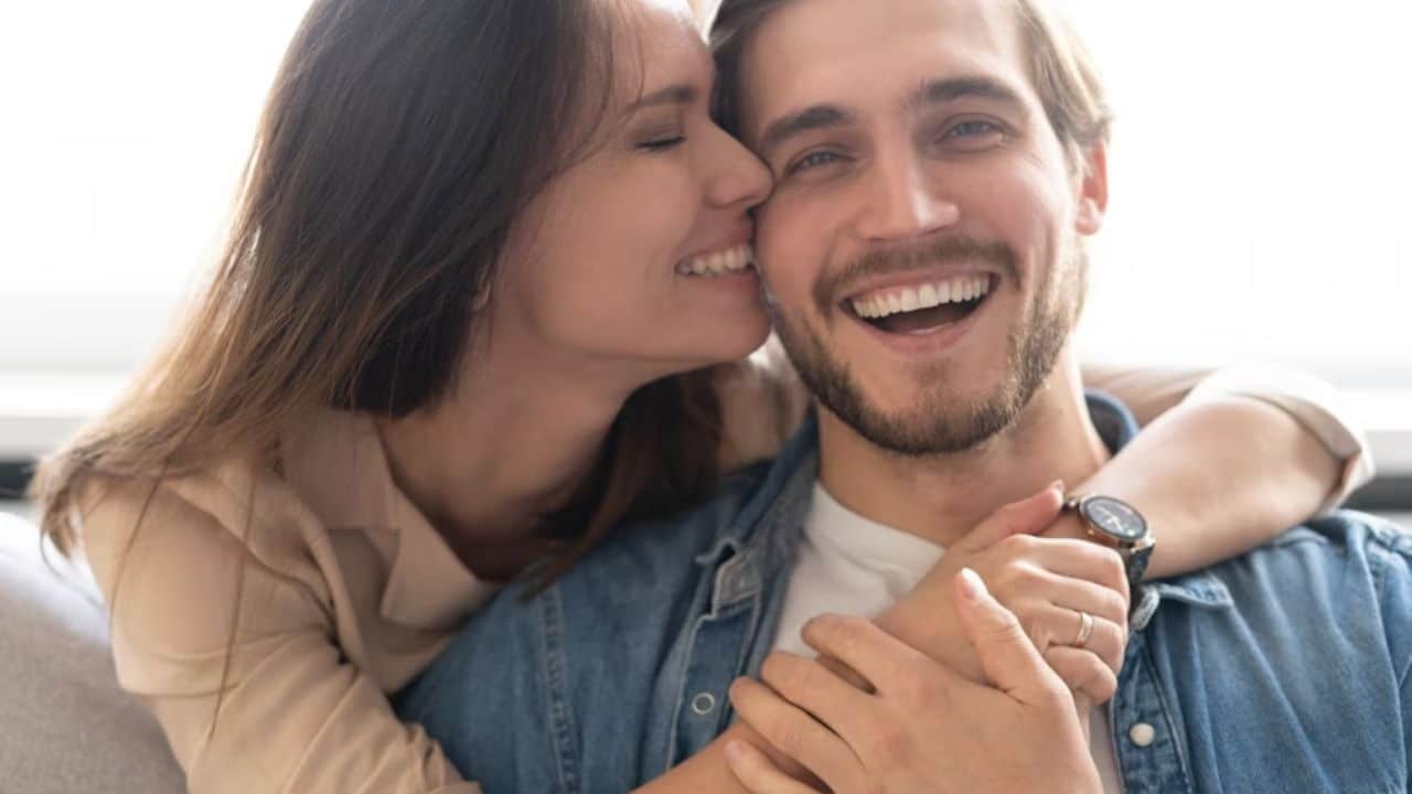 A man smiling while a woman with closed eyes folds her arms around his shoulders and leans in to kiss him on the cheek.