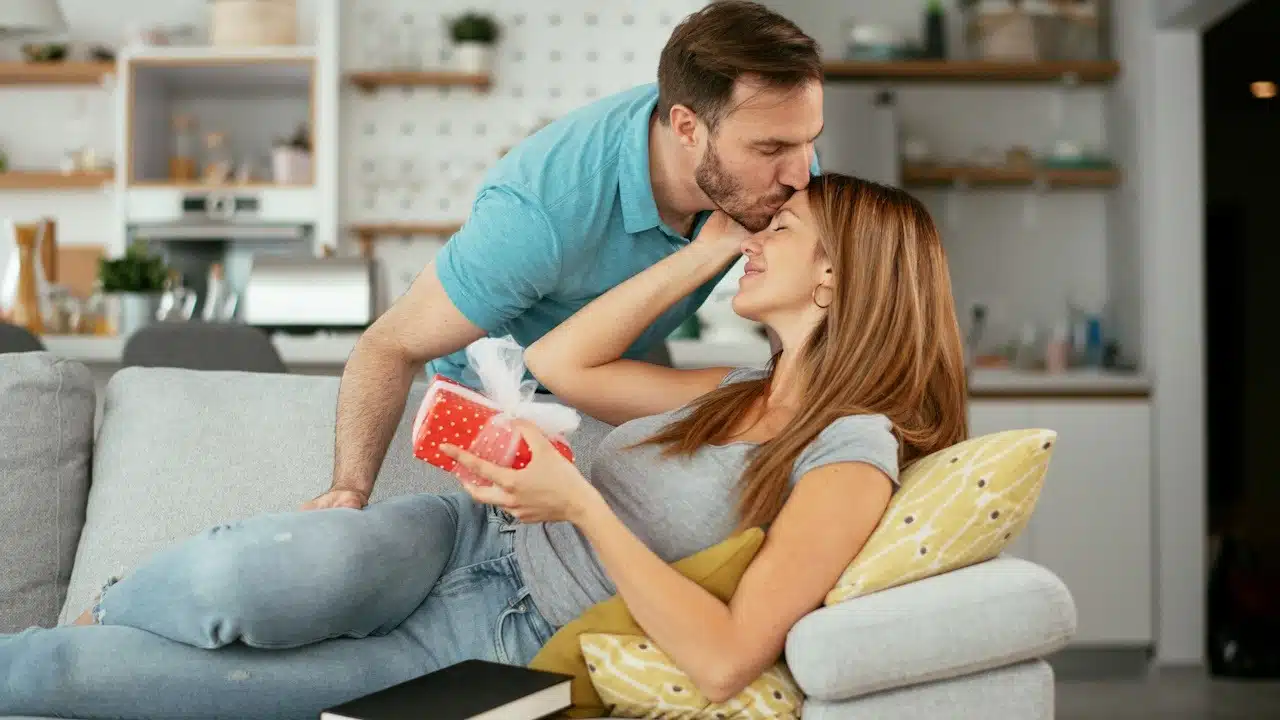 A lightly bearded man kissing the forehead of a woman who lies on a couch and holds a present.
