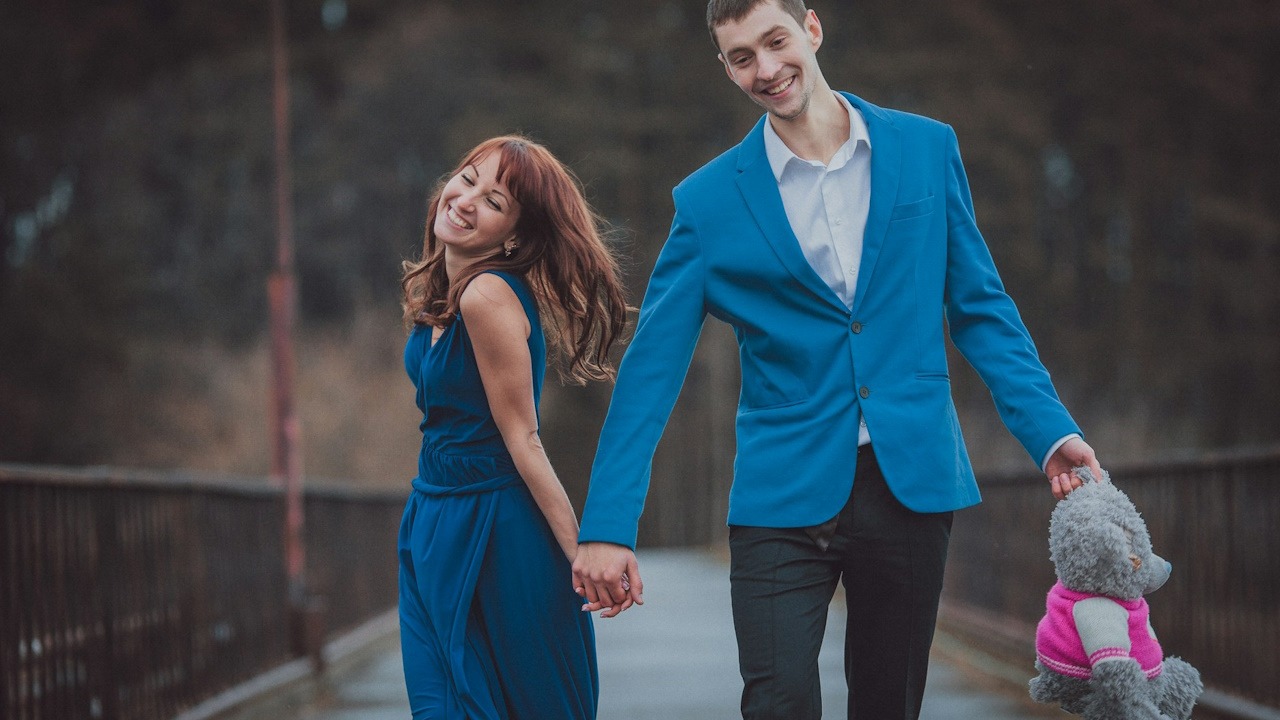 A smiling couple walking on a bridge while holding hands as the man holds a teddy bear in his free hand.