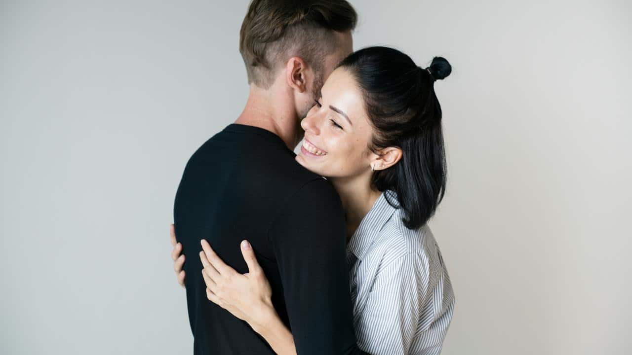 A smiling couple hugging closely against a plain background.