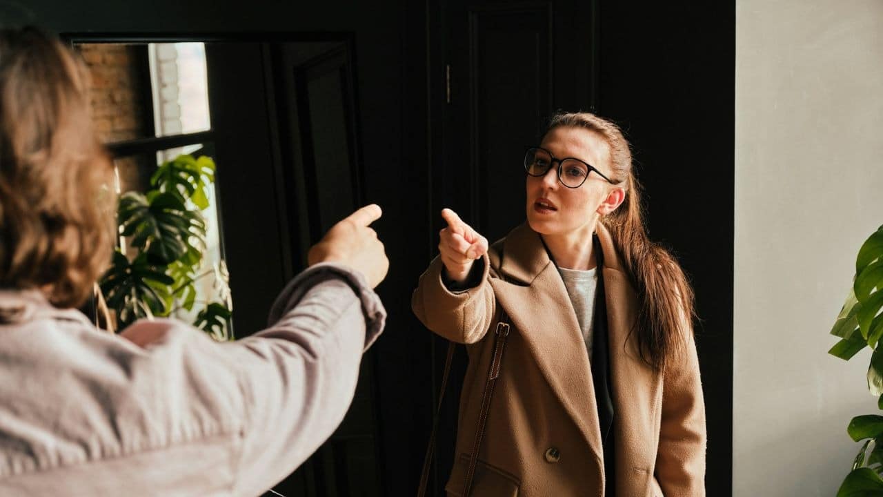 Woman in a tan coat and glasses pointing at a person facing away from the camera.