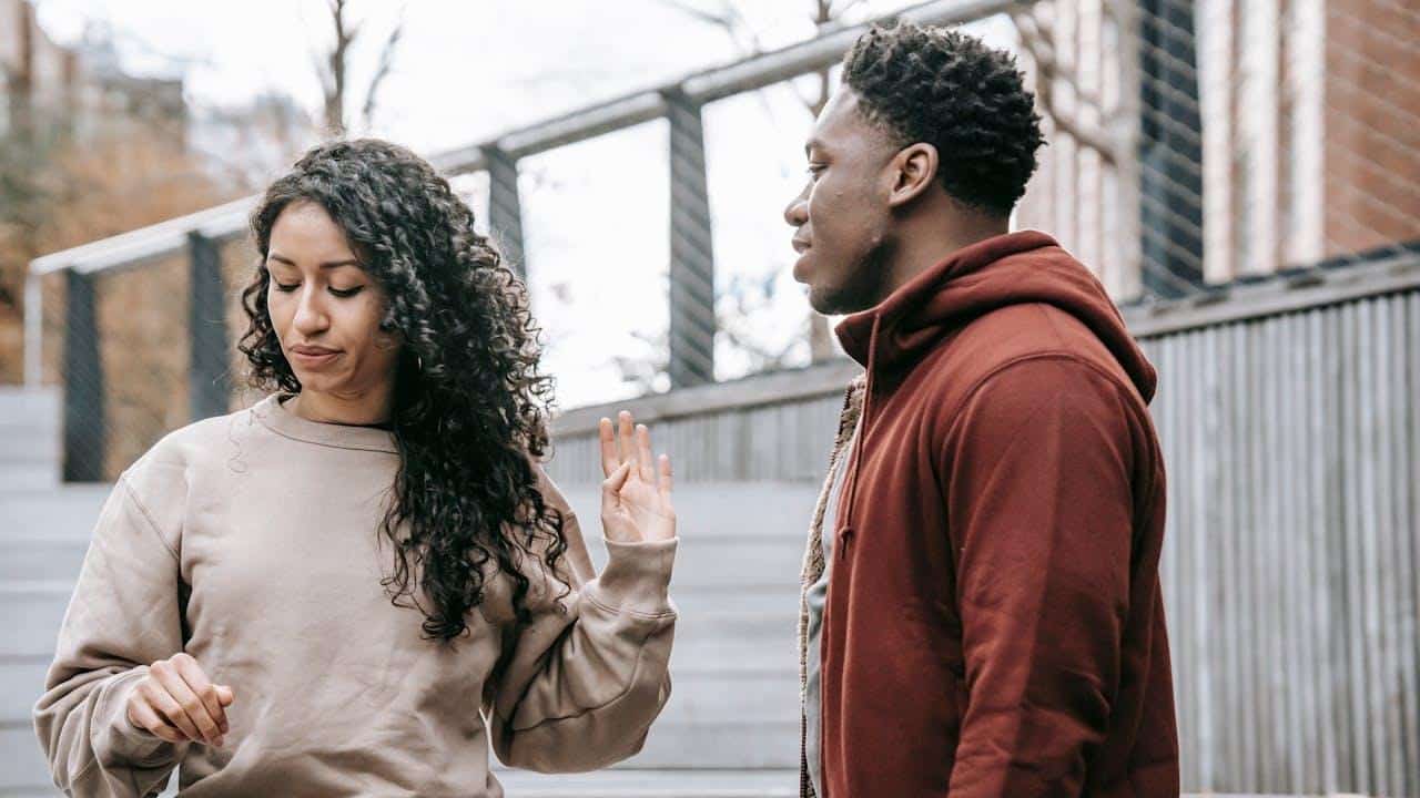 A woman with long curly hair is holding up her hand toward a man in a red hoodie outdoors.