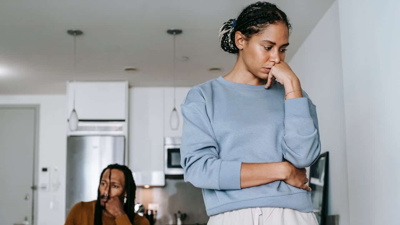 An upset woman stands in the foreground, ignoring a man sitting distressed in the background kitchen.