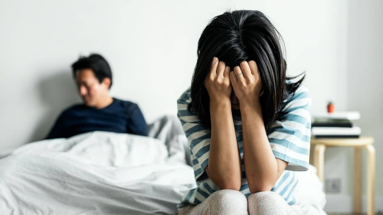 A woman sits crying with her face in her hands, a man sits behind her on a bed.