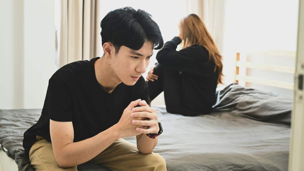A pensive man sitting on a bed, with a woman crying in the background.