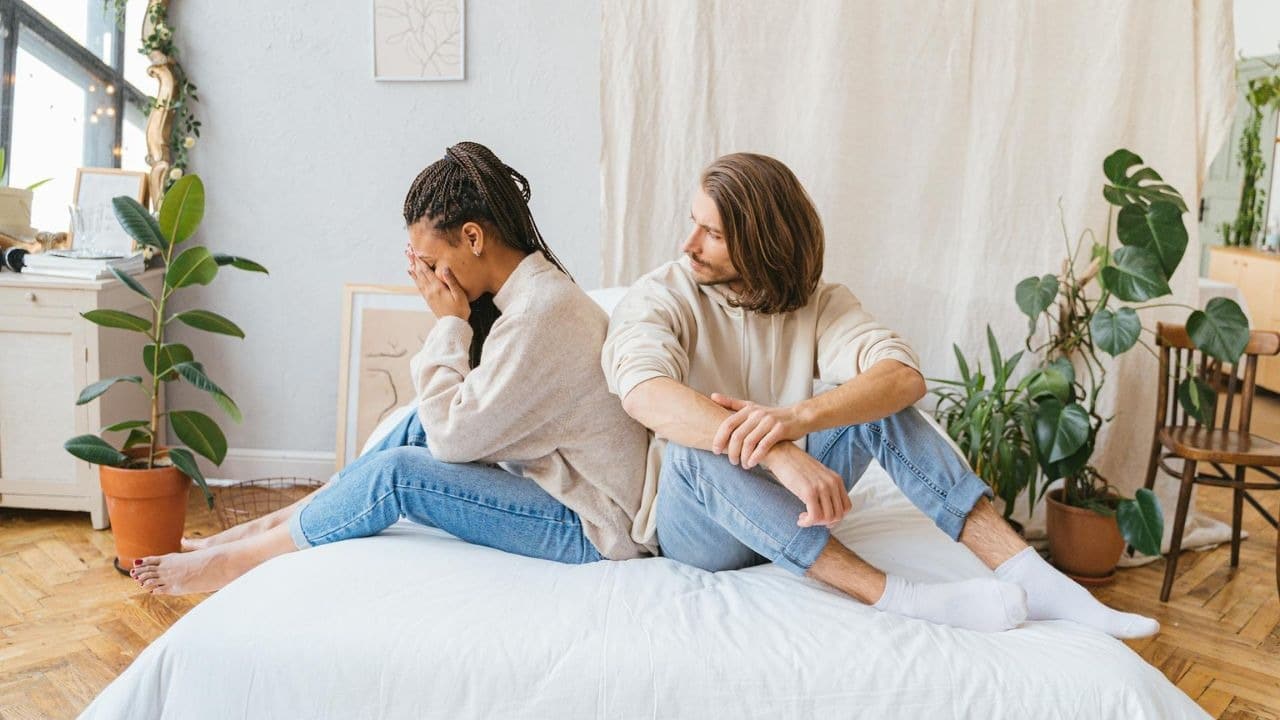 A woman sits on a bed crying into her hands while a man sits facing away.