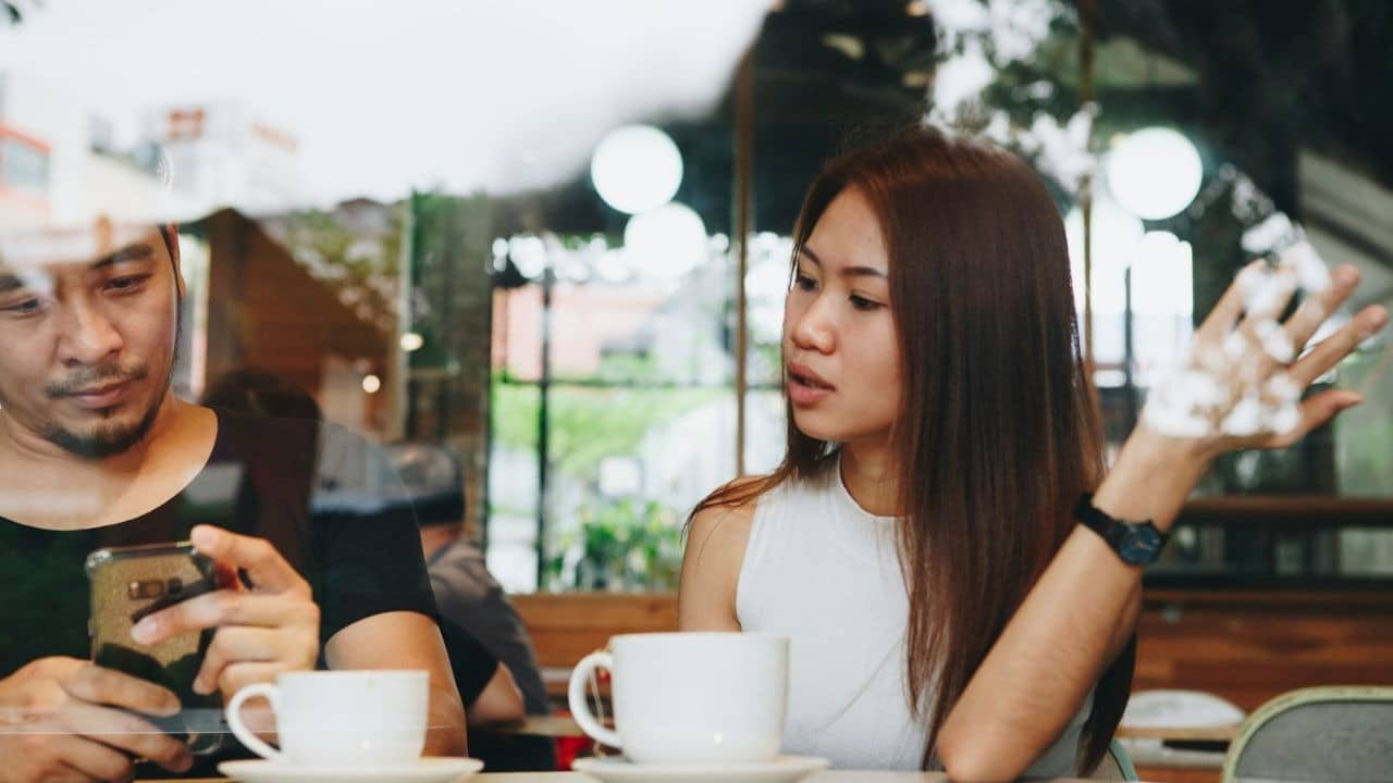 A man is looking at his phone while a woman across from him gestures and speaks