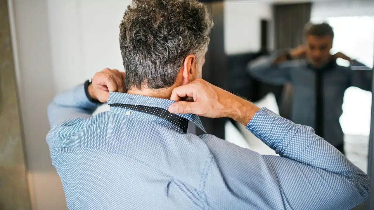 A man seen from behind adjusts his tie in front of a mirror.