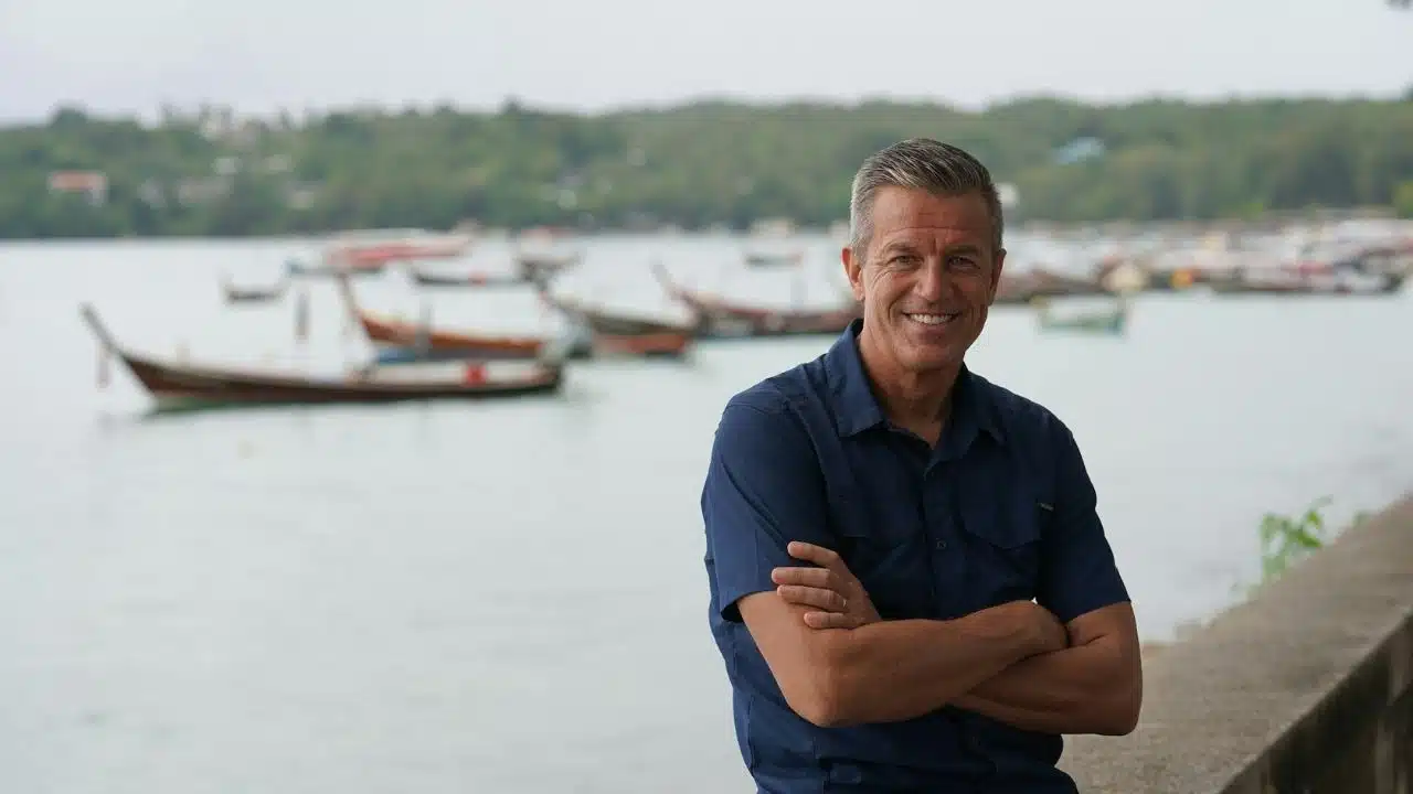 A smiling man with crossed arms stands by a body of water with boats in the background.