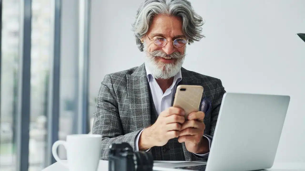 A smiling, bearded man with glasses looks at his phone while sitting at a desk with a laptop.
