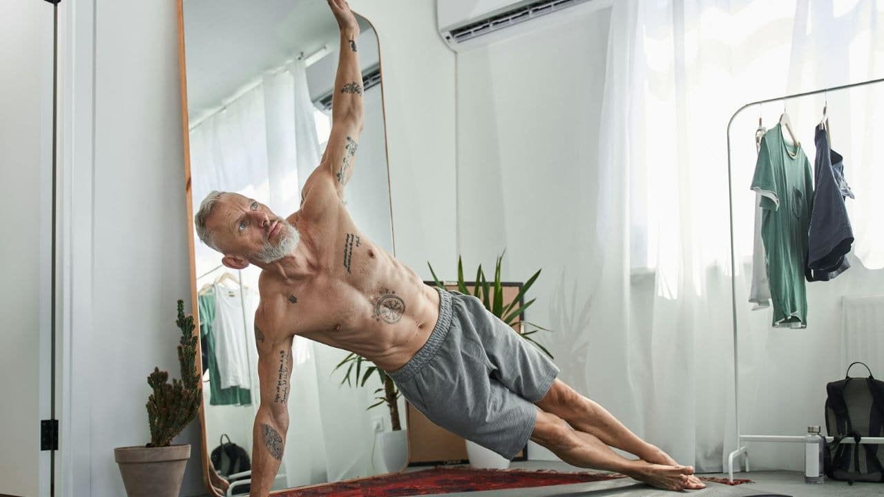 A man in a white tank top does push-ups on a wooden floor in a living room.