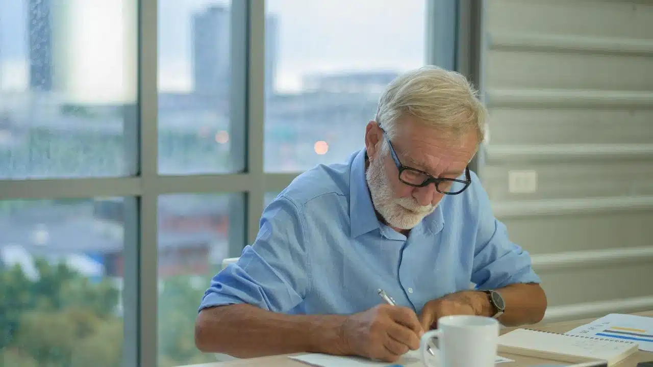 An elderly man with glasses and a beard writes at a desk next to a window.