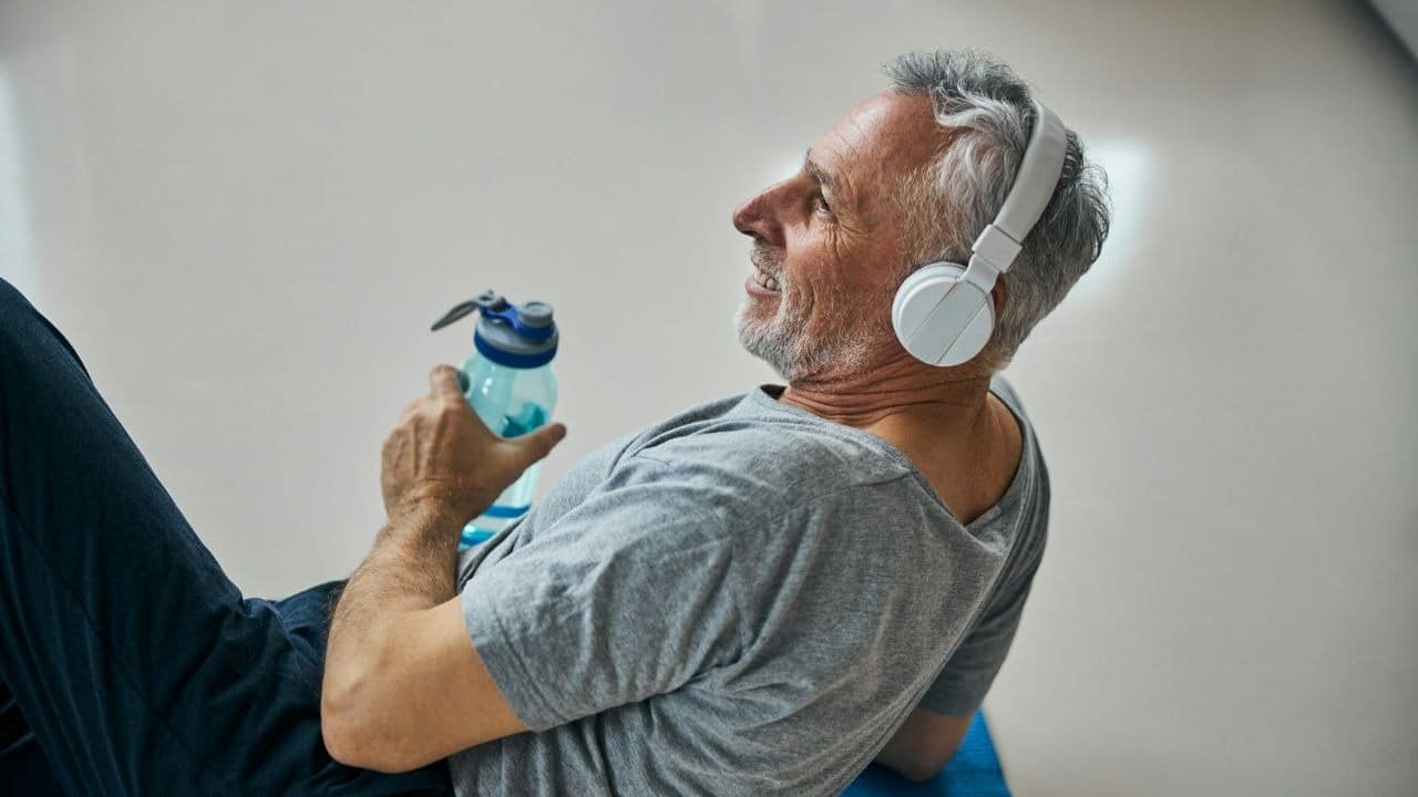 A smiling, grey-haired man with headphones lies down, holding a water bottle.