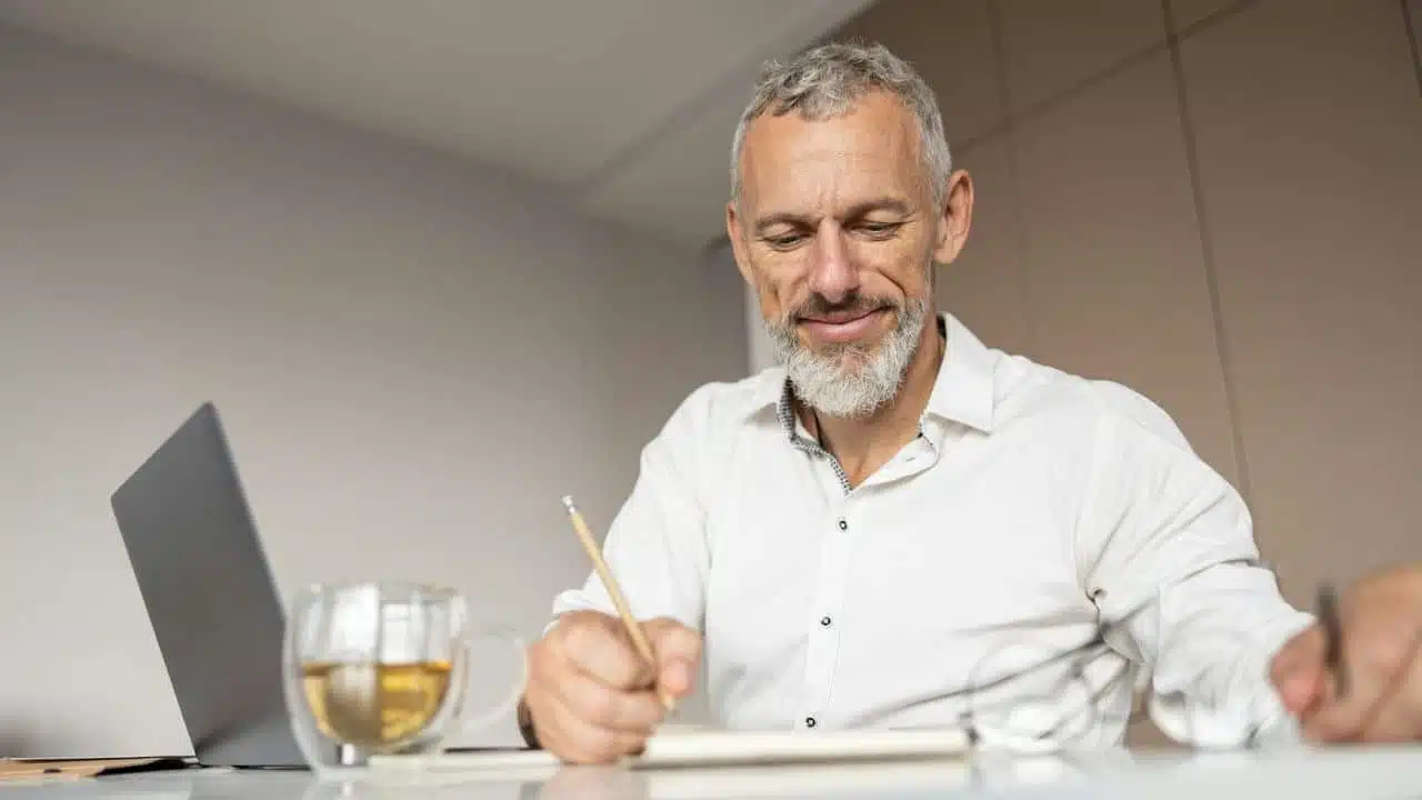 A smiling man with grey hair and beard stands against a brick wall.