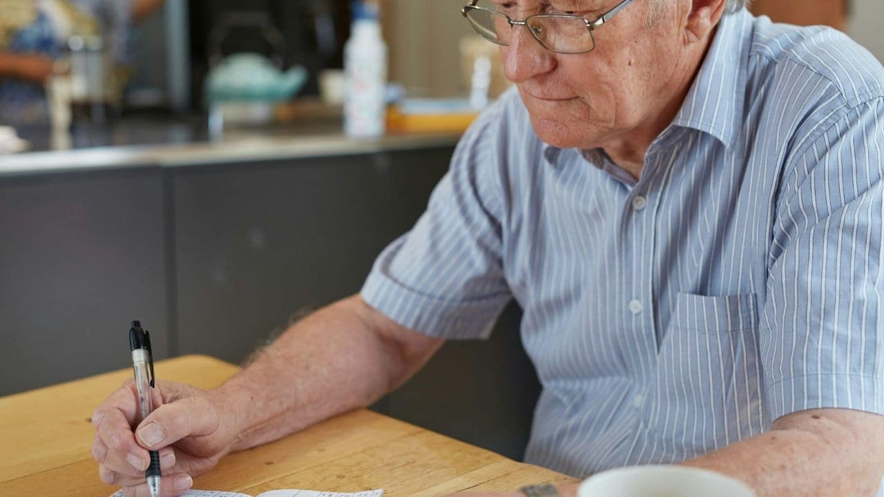 A man with glasses writes in a notebook at a wooden table, holding a pen.