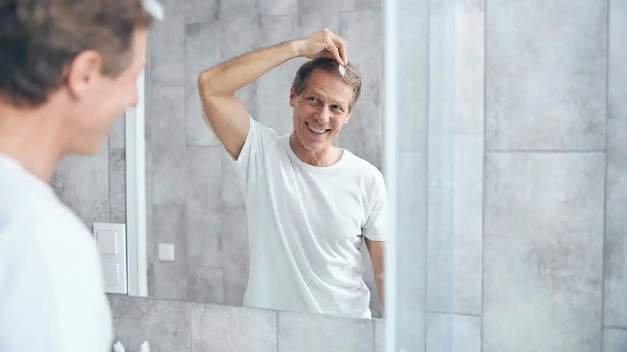 A smiling man in a white t-shirt looks in a mirror, touching his hair.