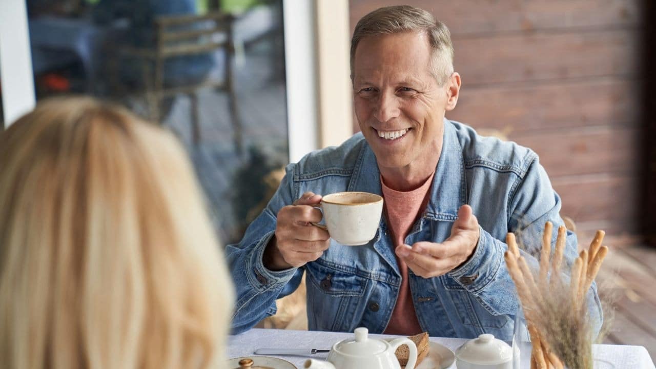 A smiling man in a denim jacket holds a cup and gestures, looking at someone across a table.