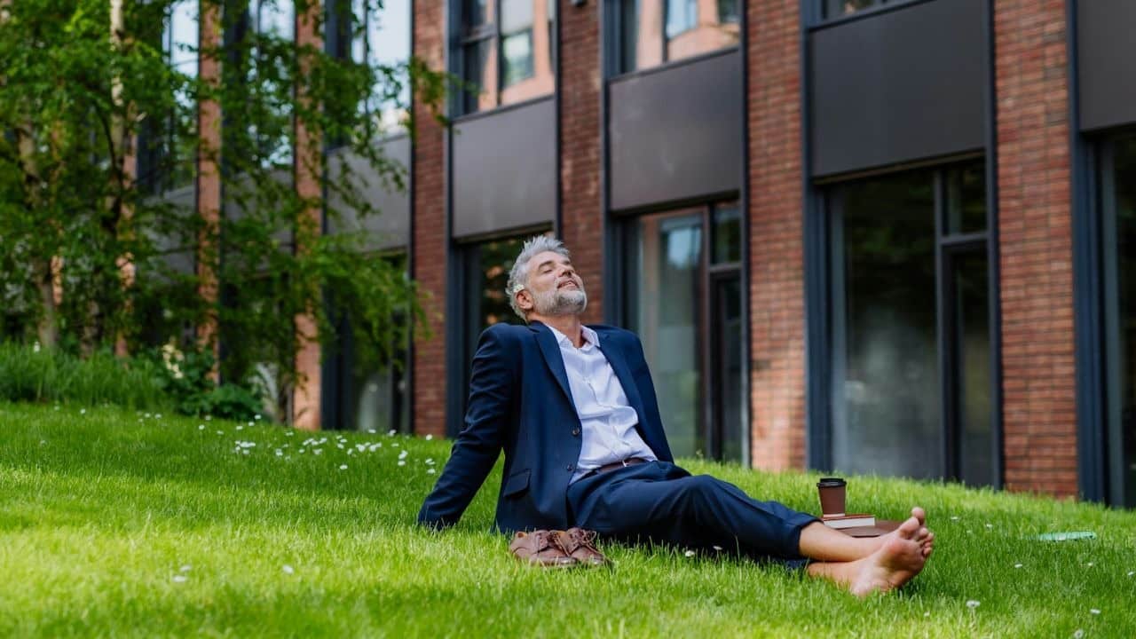 A relaxed, grey-haired man in a suit lies barefoot on a grassy hill, eyes closed.