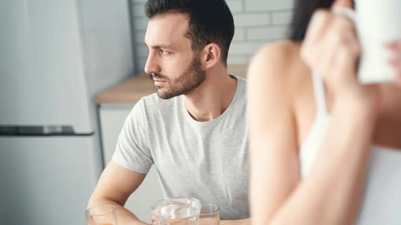 A man with a beard looks away from a woman, with drinks on a table.