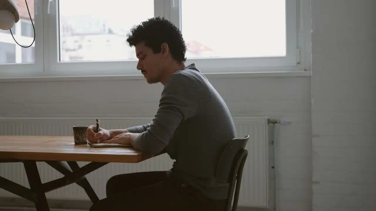 A man with curly hair and a mustache sits at a wooden table, writing in a notebook.