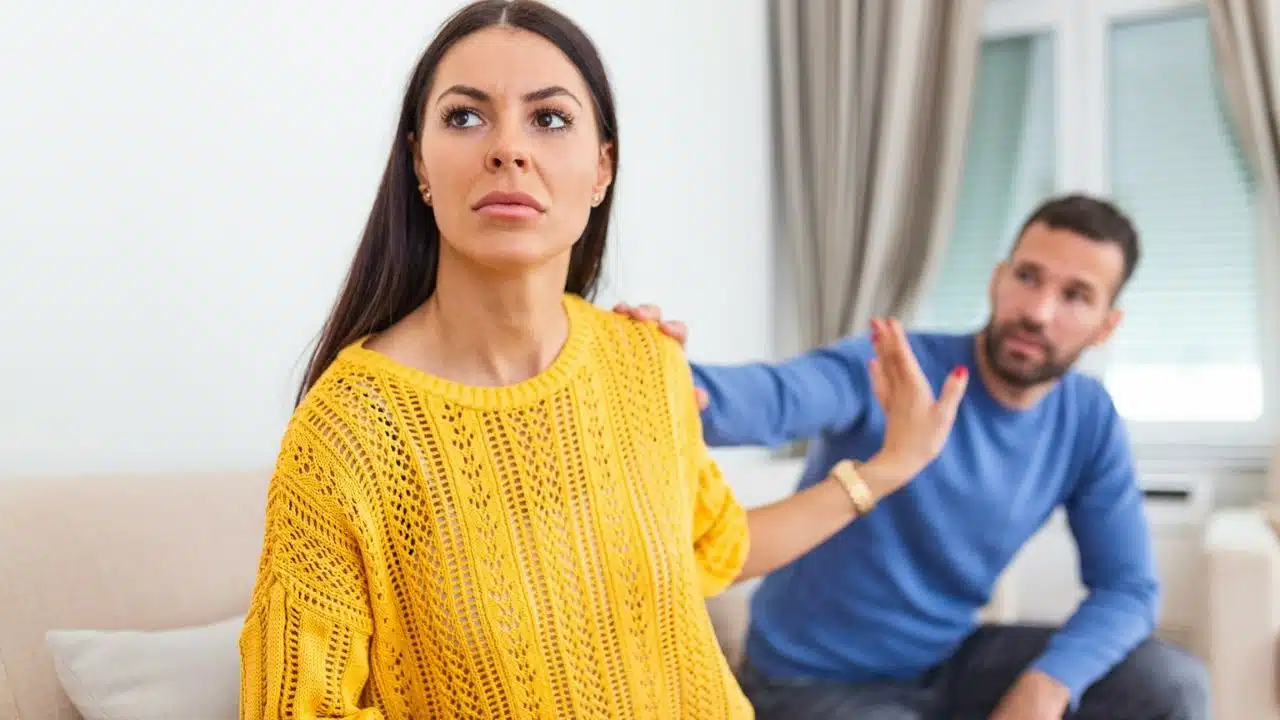 A woman in a yellow sweater looks away with a sad expression as a man gestures behind her.