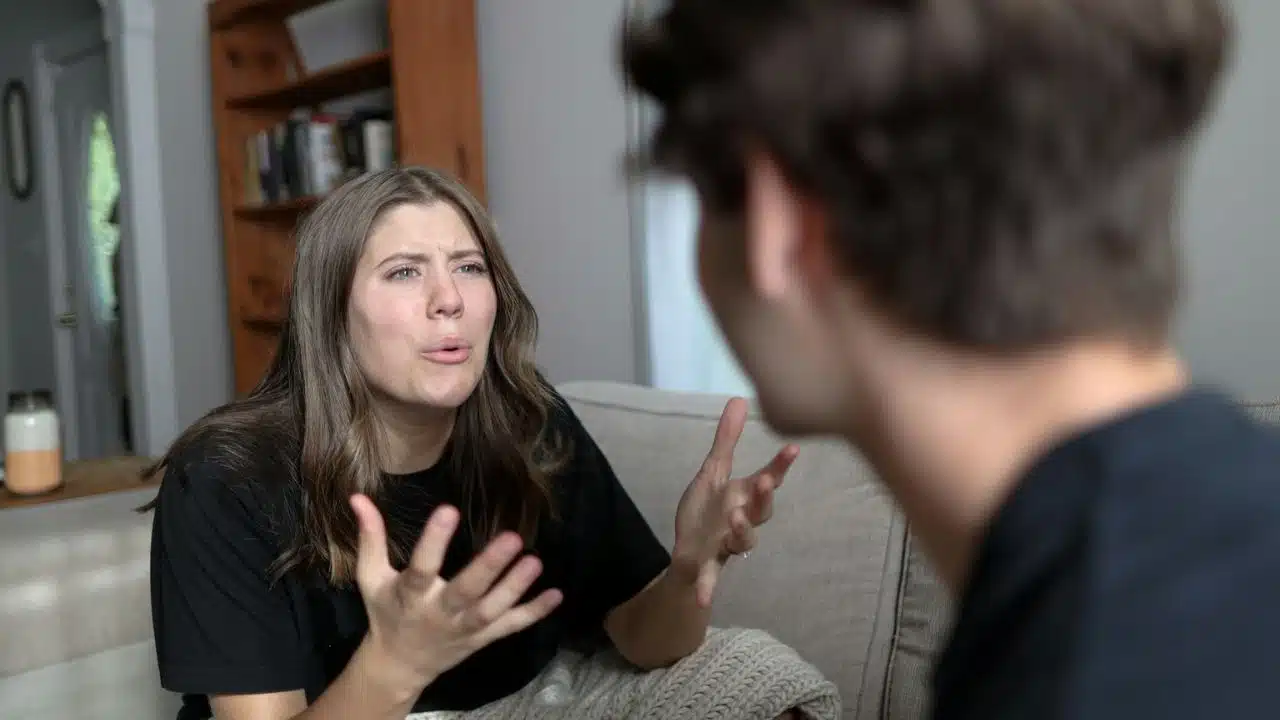 A woman with long hair gestures emphatically while speaking to a man facing away.
