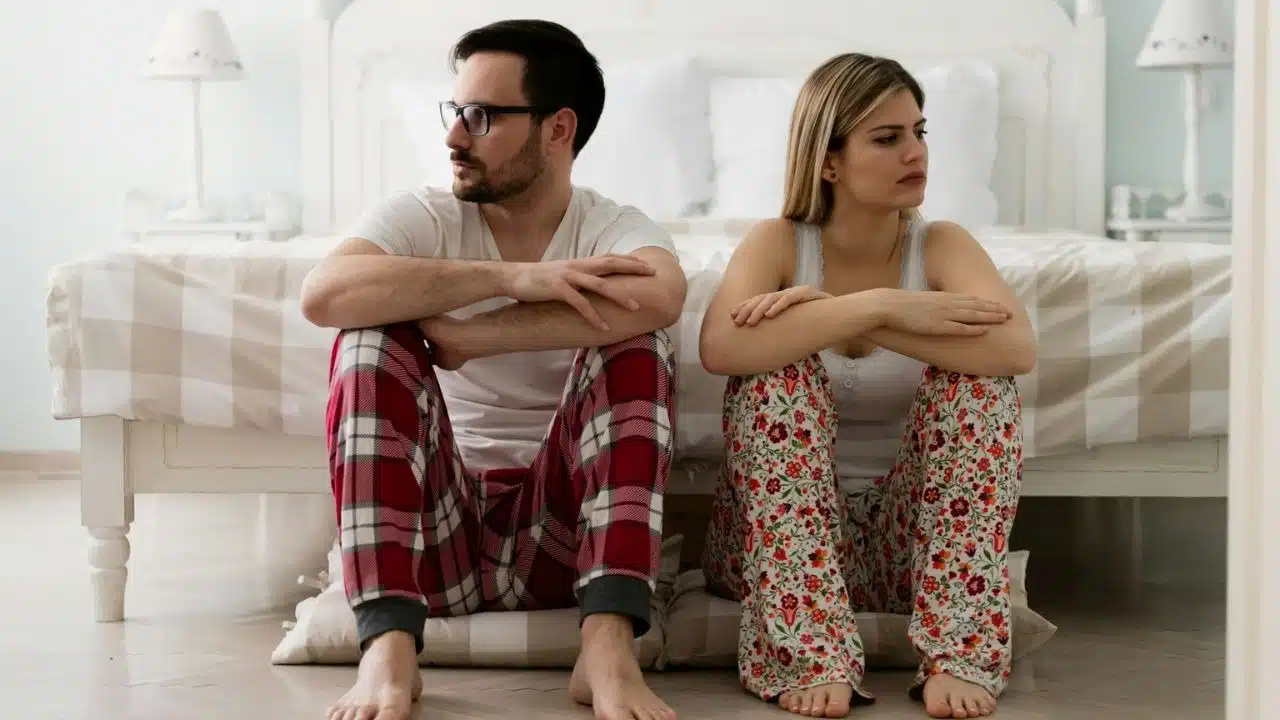 A man and a woman in pajamas sit on the floor, looking away from each other.