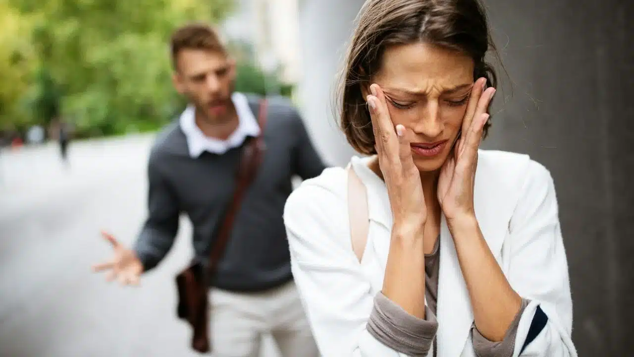 A distressed woman covers her ears as a man shouts and gestures behind her.